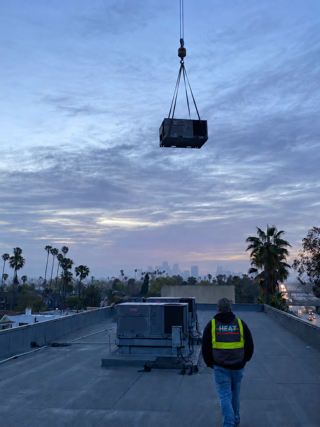 Crane lowering a rooftop HVAC unit into place at sunset with the Los Angeles skyline in the background